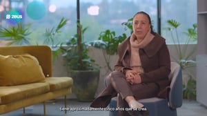 Persona sentada en un sillón dentro de una oficina moderna, con plantas y ventanales al fondo. En pantalla aparece el logotipo de Zeus y un subtítulo. Escena grabada en interior, estilo entrevista corporativa.